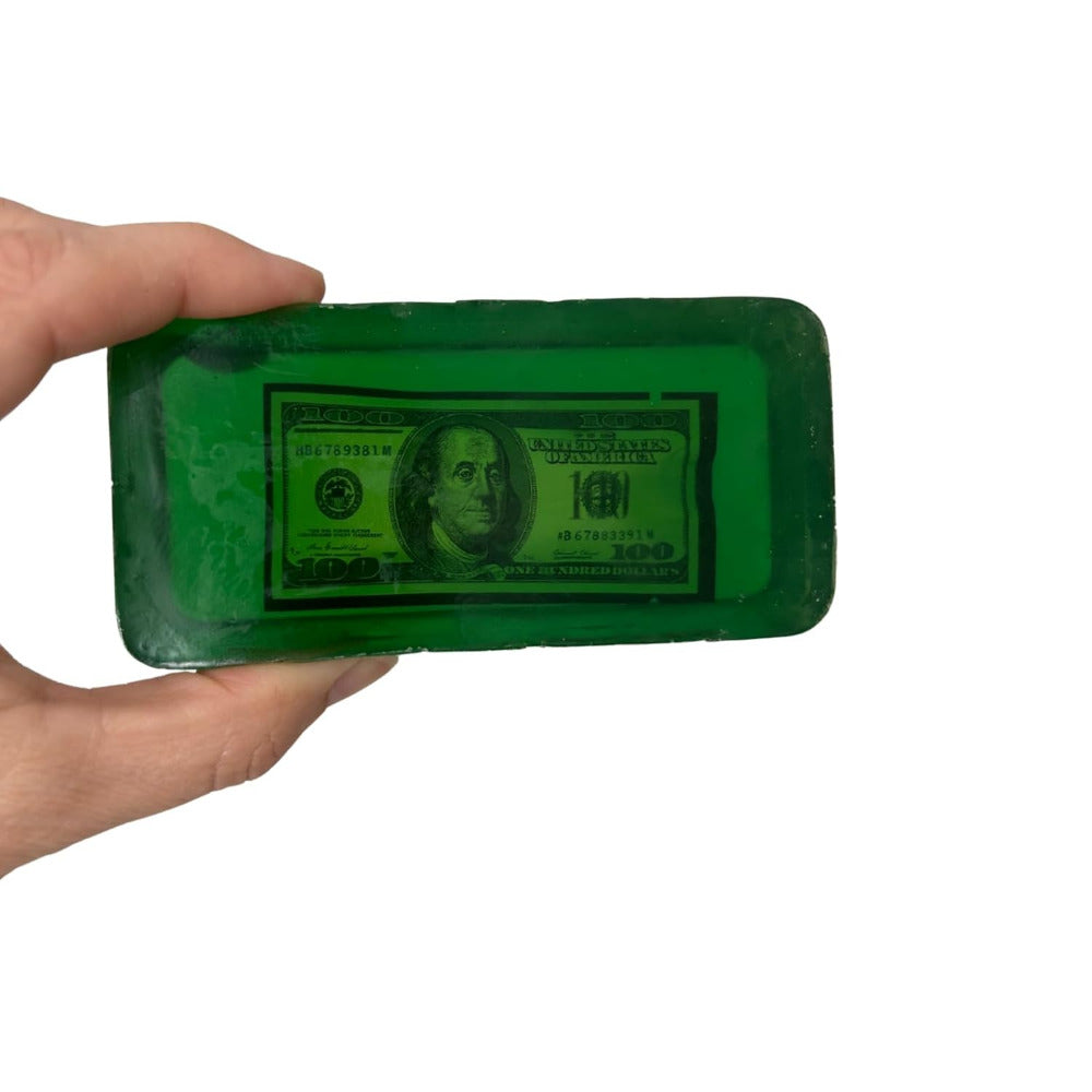 Green bar of money soap with a dollar bill design held by a hand on a white background